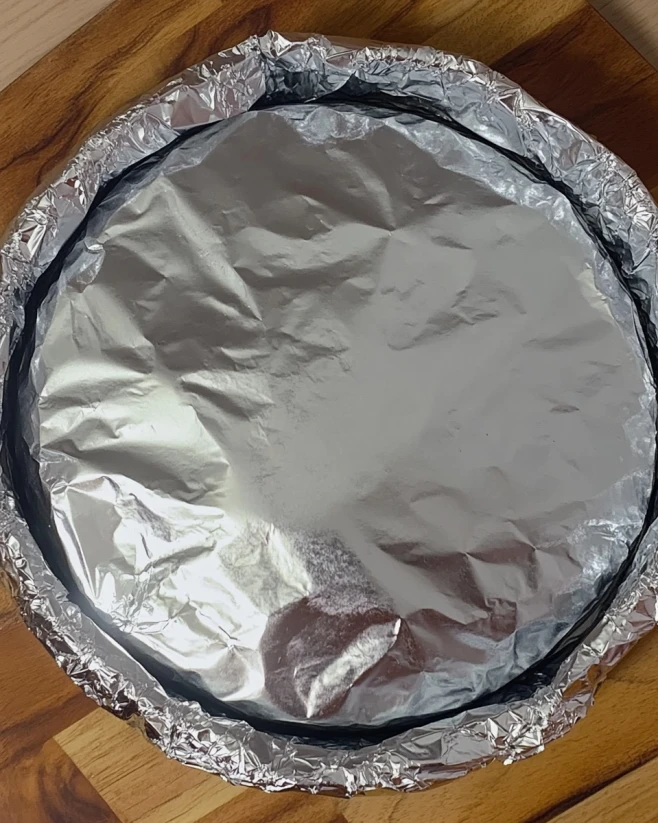 Pistachio Cottage Cheese Cheesecake Ready for Baking Foil-covered springform pan prepared for baking pistachio cottage cheese cheesecake on a wooden surface