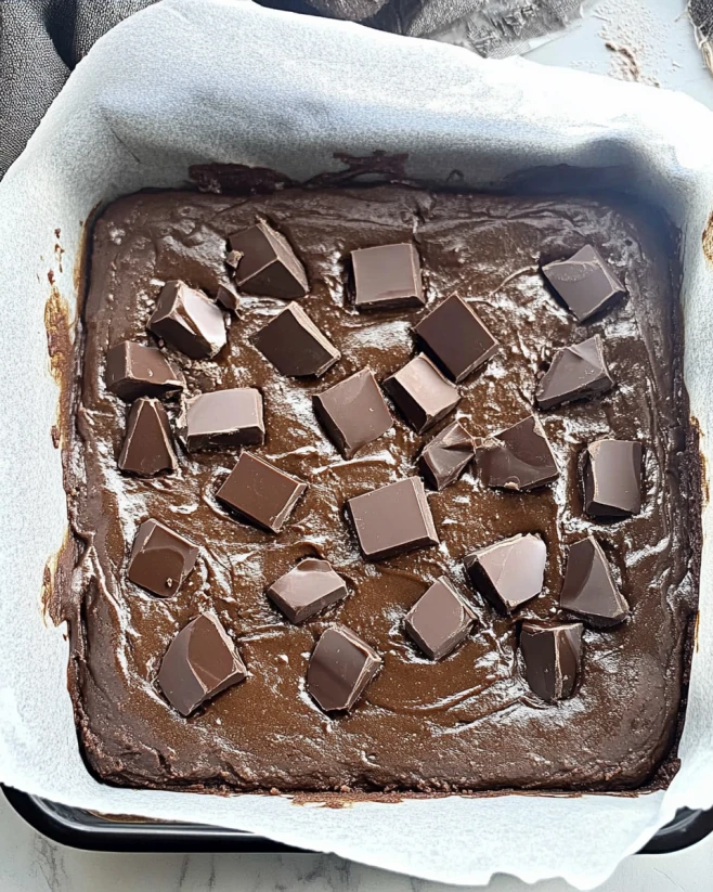 Cottage Cheese Brownie Batter Topped with Chocolate Chunks Unbaked brownie batter in a parchment-lined pan with chocolate chunks on top, made with cottage cheese.